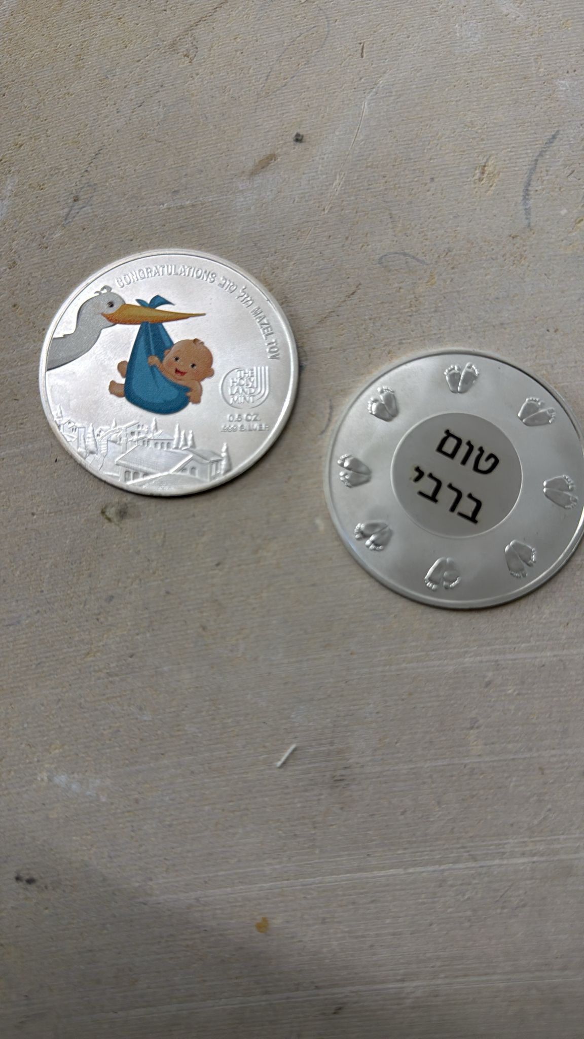חריטה על מטבע כסף עם השם טום ברבי לצד מטבע מזל טוב מאויר עם חסידה ותינוק