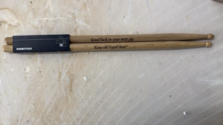 חריטה על זוג מקלות תופים מעץ עם הכיתוב Good luck in your next gig ו Keep the hyper beat