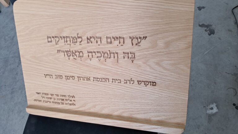 חריטה על סטנדר לסידור מעץ אלון עם הפסוק עץ חיים היא למחזיקים בה והקדשה לרב אהרון סימן טוב